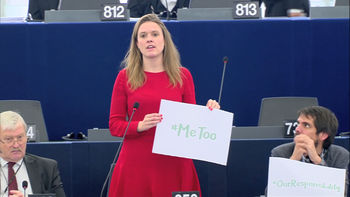 En kvinne i rød kjole står i en sal og holder et skilt med teksten "Me Too"