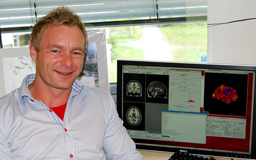 En smilende mann (professor Anders Fjell) sitter foran en dataskjerm.