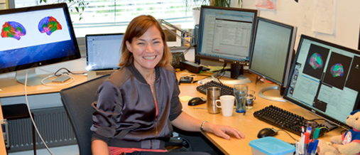 En smilende kvinne (professor Kristine Walhovd) sitter ved en kontorpult med flere skjermer.