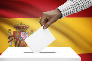 A hand placing a ballot in a box in front of the Spanish flag.