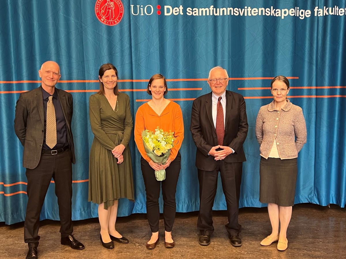 Five smiling people in front of a blue curtain. The woman in the middle (Anke Schwarzkopf) is holding a boquet of flowers.