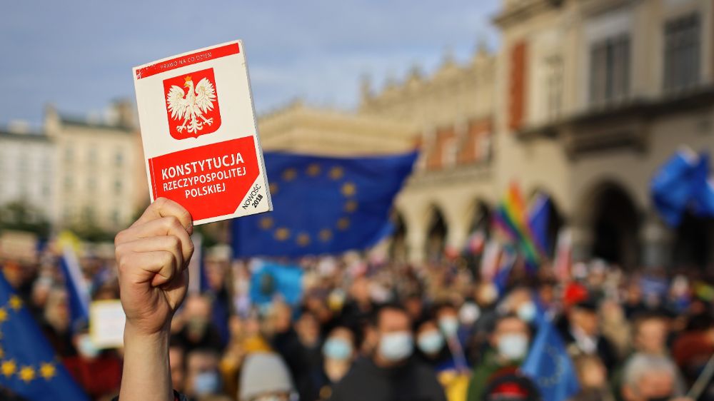 En hånd holder opp en utgave av den polske grunnloven. Demonstrasjoner med EU-flagget i bakgrunnen.