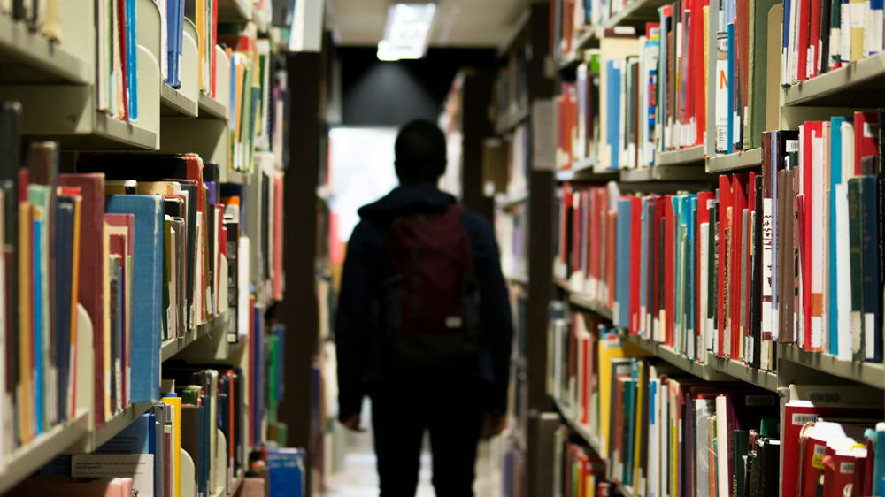 svartkledd person står mellom bokhyller i et bibliotek