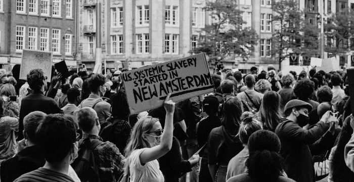 black and white photo of a protest, one woman is holding a sign reading "US systemic slavery started in New Amsterdam"