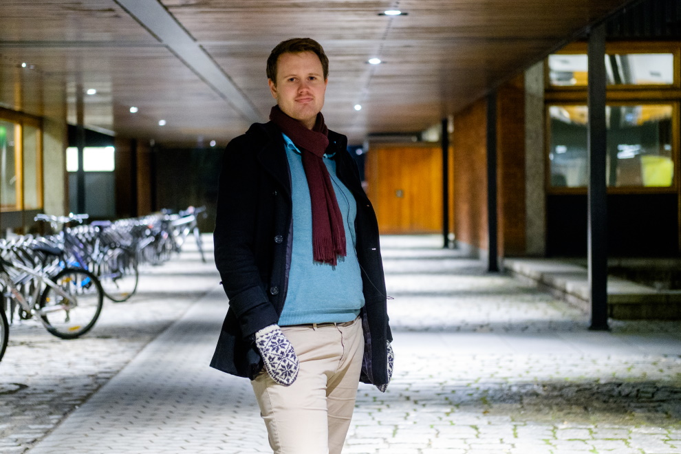 Ung mann med mørkt hår, mørkerødt skjerf og selvbuvotter står smilende foran sykkelstativ