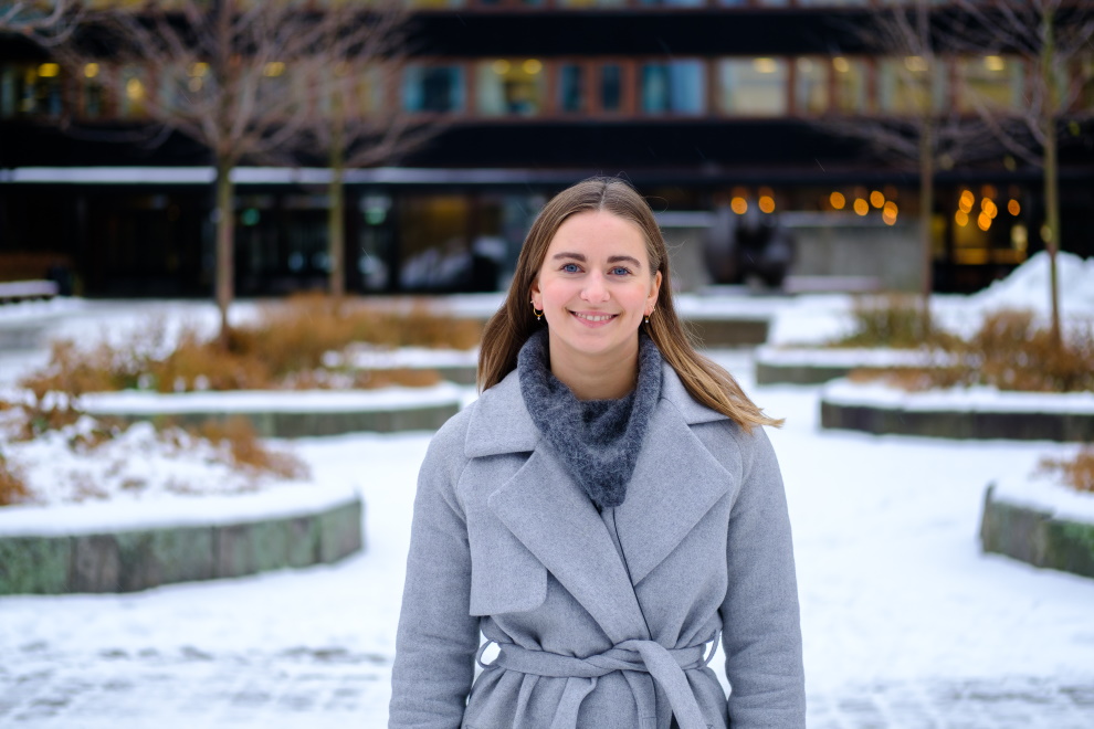 Ung kvinne med brunt hår og grå kåpe står ute og smiler