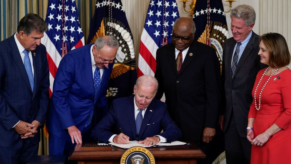 Bilde av Joe Biden sittende ved et skrivebord signerer klimaavtale med andre politikere stående rundt bordet