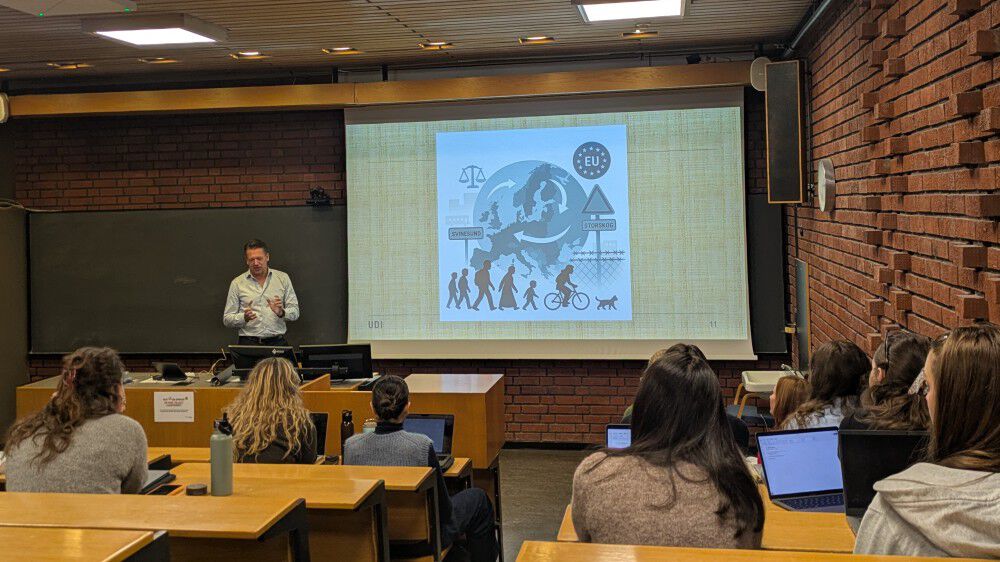 Bildet viser UDI-direktør Snorre Sæther som holder en presentasjon i et klassisk Blindern-auditorium – det vil si vegger i rød-oransje teglstein, grønn tavle og fastmonterte bord og stoler i lys tre. Man ser også studentene som følger foredraget bakfra. Mange har PC’er og drikkeflasker foran seg.