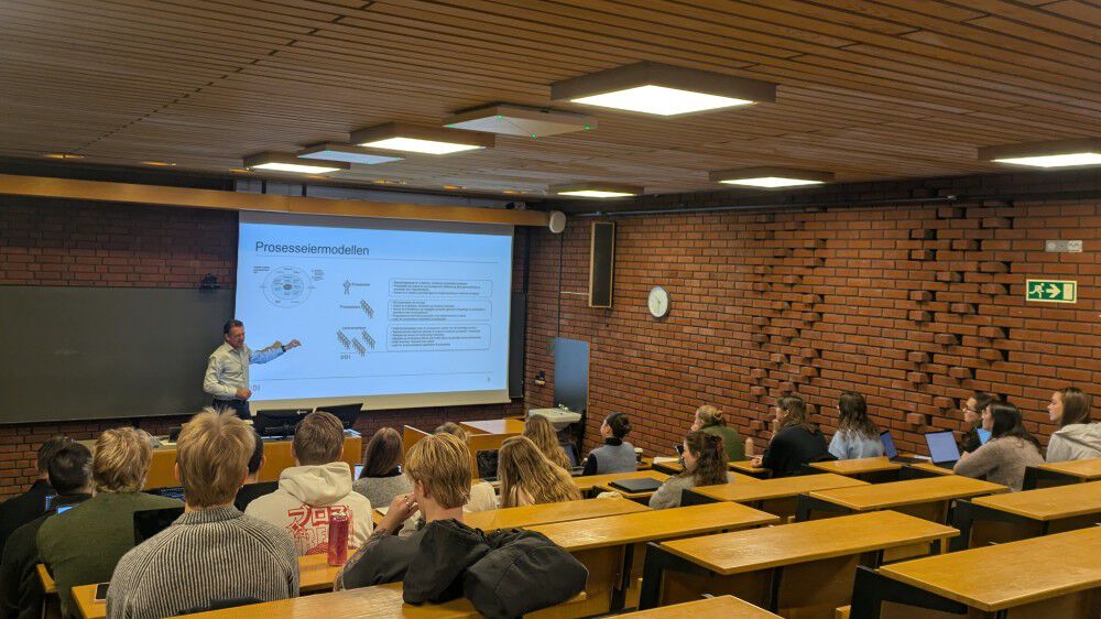 Bildet viser UDI-direktør Snorre Sæther som holder en presentasjon i et klassisk Blindern-auditorium – det vil si vegger i rød-oransje teglstein, grønn tavle og fastmonterte bord og stoler i lys tre. Man ser også studentene som følger foredraget bakfra. Mange har PC’er og drikkeflasker foran seg.