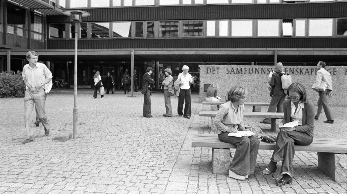 Svart-hvitt fotografi av studenter utenfor Eilert Sundts hus, noen gående, andre sittende på benker.