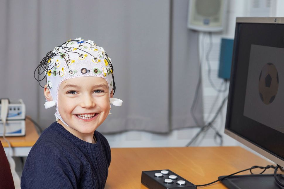 Image contains a smiling child, wearing a cap  for research purposes, sitting at a desk.