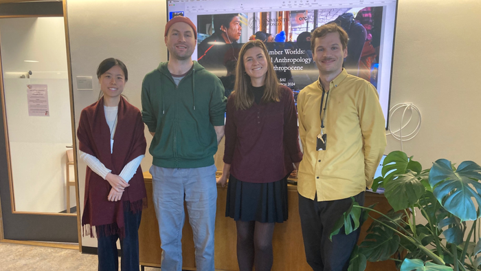 Four persons standing side by side and smiling in front of a large screen with a power point presentation on it.