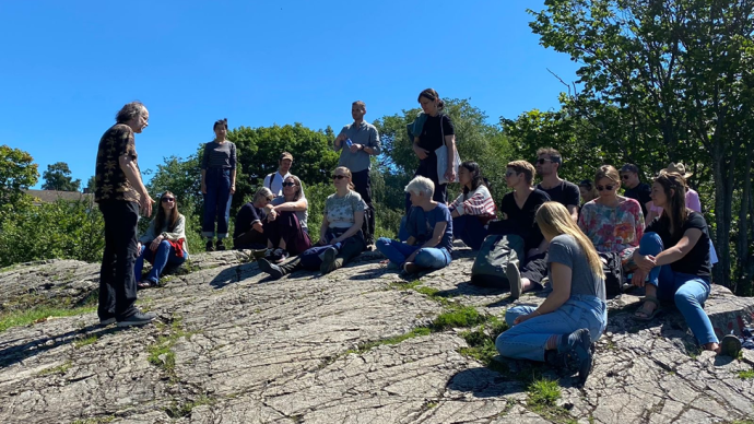 A person talking to a group of people sat around him in a semicircle, three people are also standing, they are outside sitting on a flat expanse of rock, trees are in the background and the sky is free of clouds.