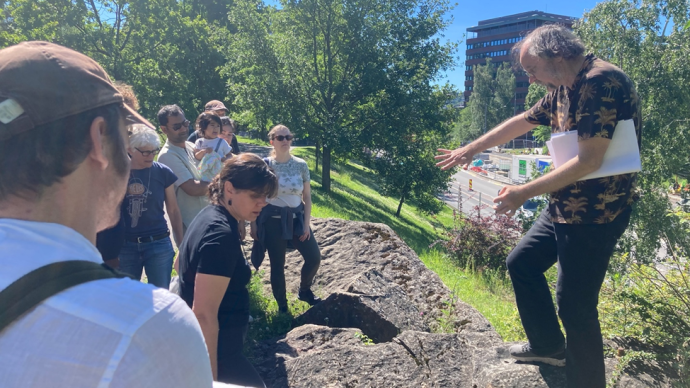 A group of people standing around a man gesticulating toward a piece of rock that the people are standing on, while carrying some papers in the crook of his arm.
