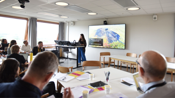 A person presenting in front of a tv-screen with a powerpoint presentation open, we see some of the other people in the room sitting and listening as well