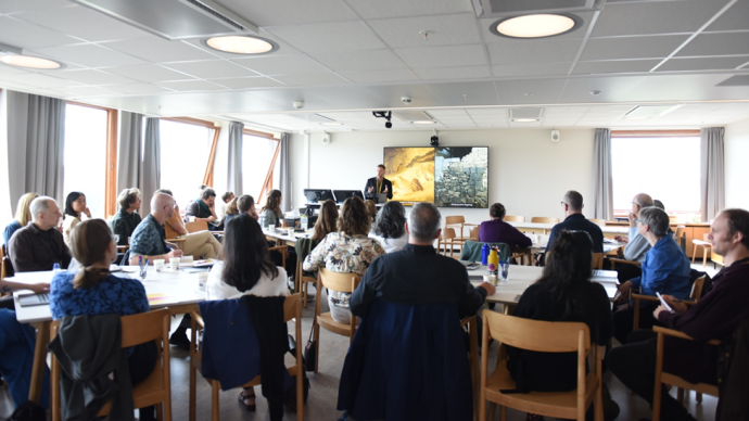 A person presenting in front of a tv-screen with a powerpoint presentation open, we see many people in the room sitting and listening as well