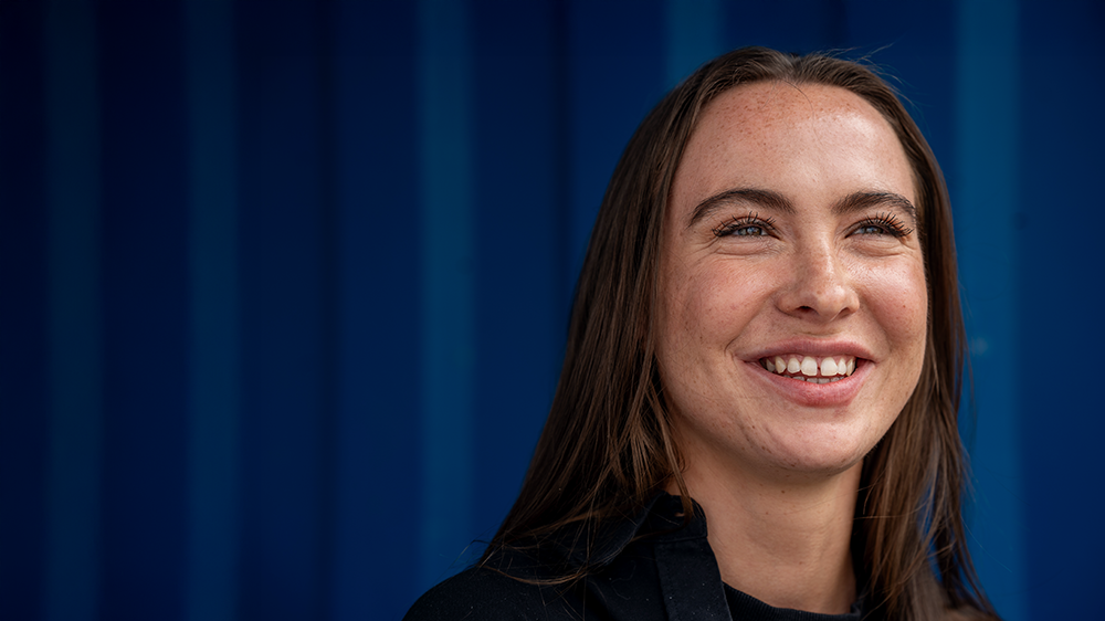 Lina, som har en bachelor i utviklingsstudier og bærekraft fra UiO, ser ut i lufta og smiler mot en blå bakgrunn