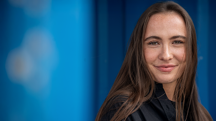 Lina, som har en bachelor i utviklingsstudier og bærekraft fra UiO, ser inn i kamera foran en blå vegg